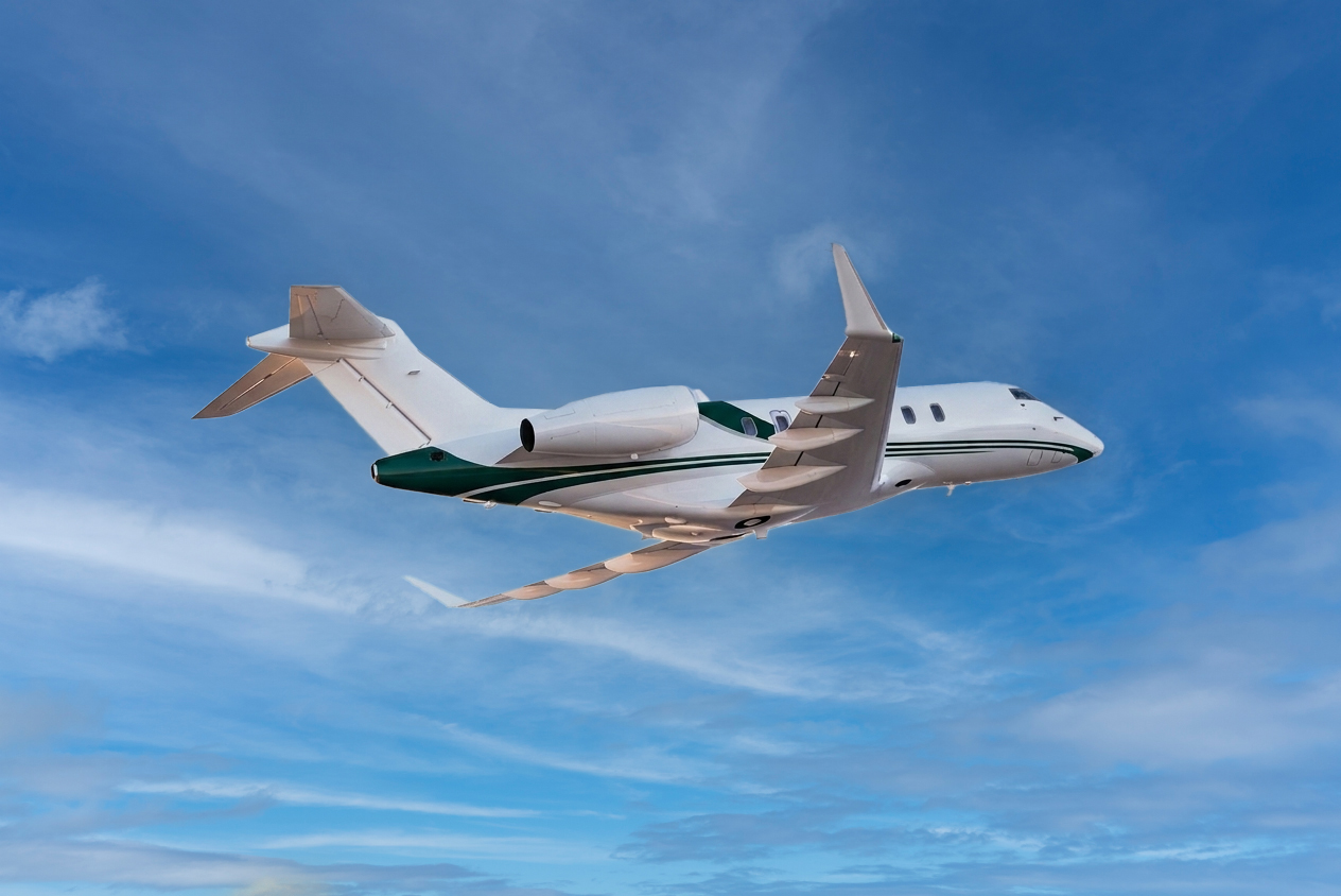 White business jet with dark green accents flying above the clouds against a bright blue sky, viewed from below in mid-flight.