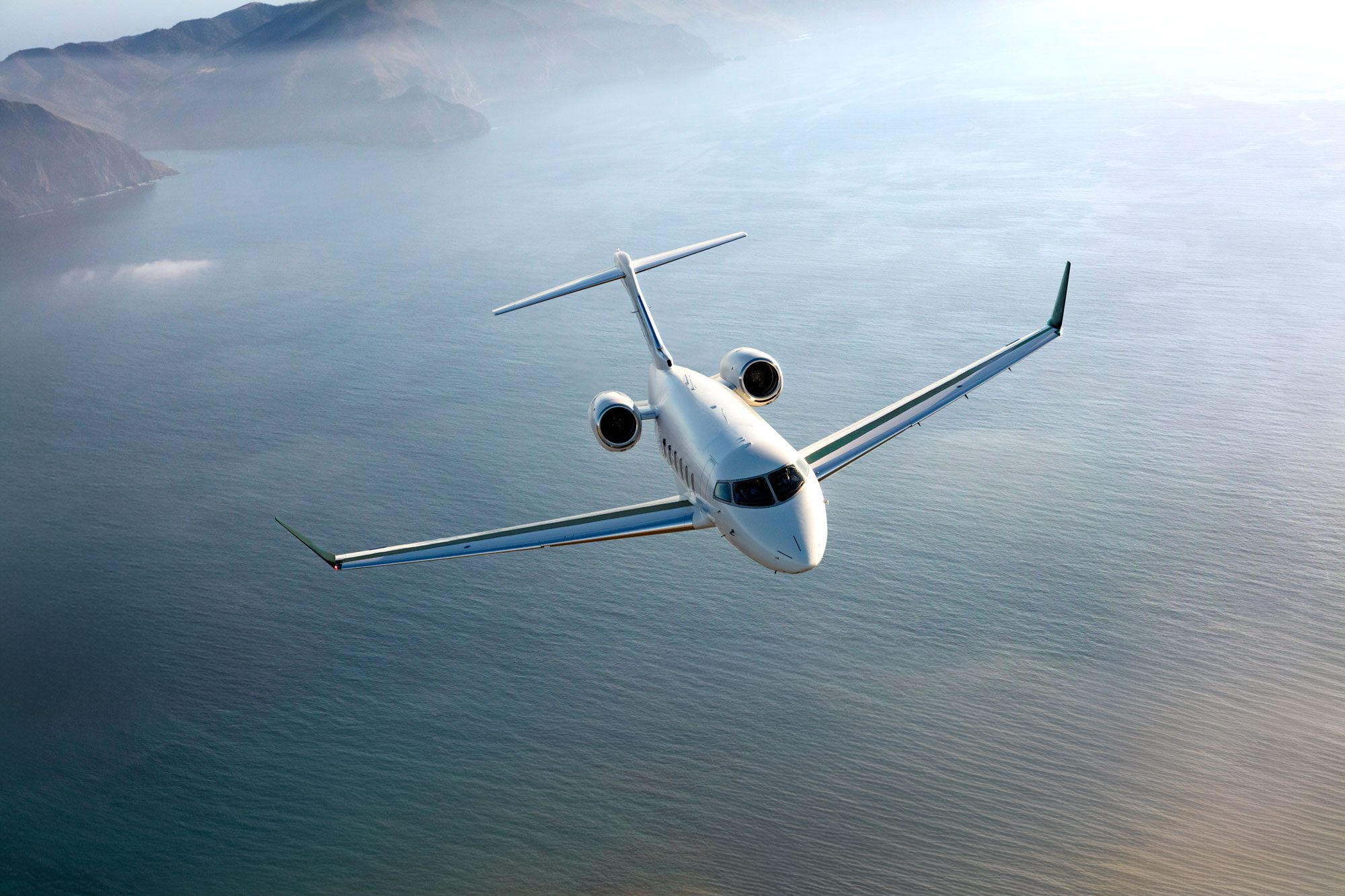 Bombardier Challenger 650 aircraft in flight over a coastal landscape.