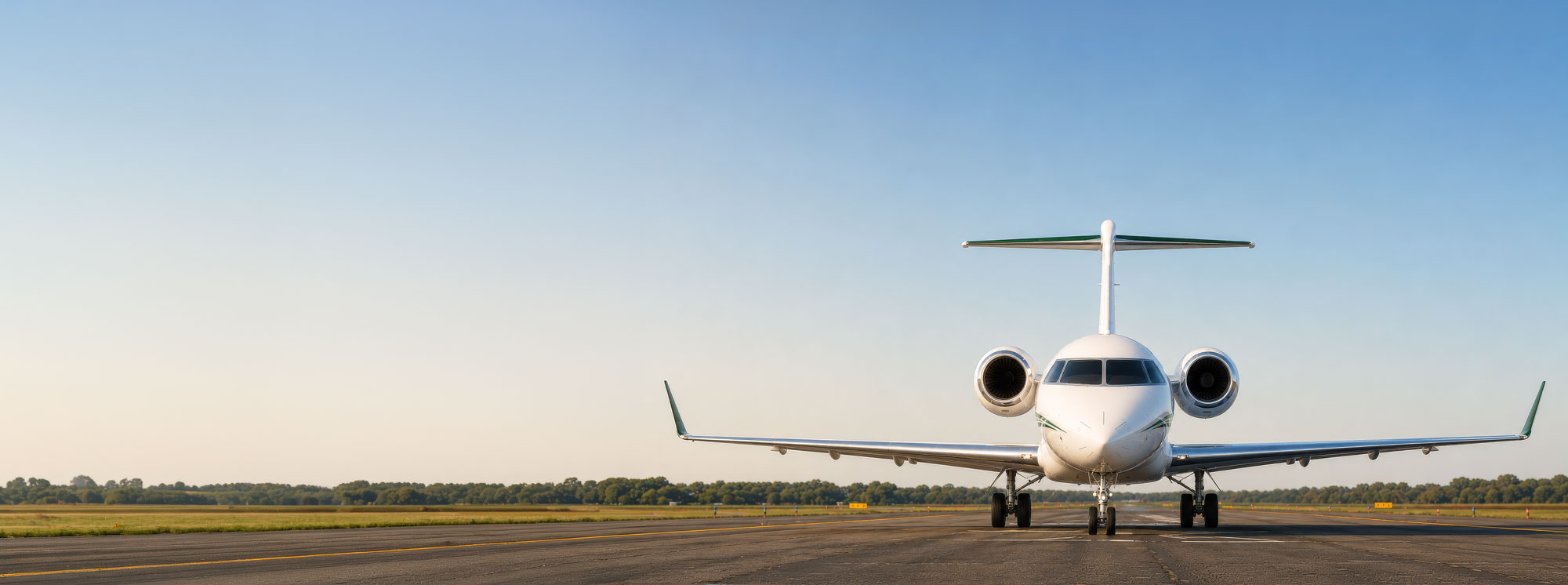 Front view of a white Bombardier Challenger 3500 business jet on a runway in warm morning sunlight, featuring dark green accents, rear-mounted engines, distinctive winglets, and detailed wing surfaces beneath a clear blue sky.