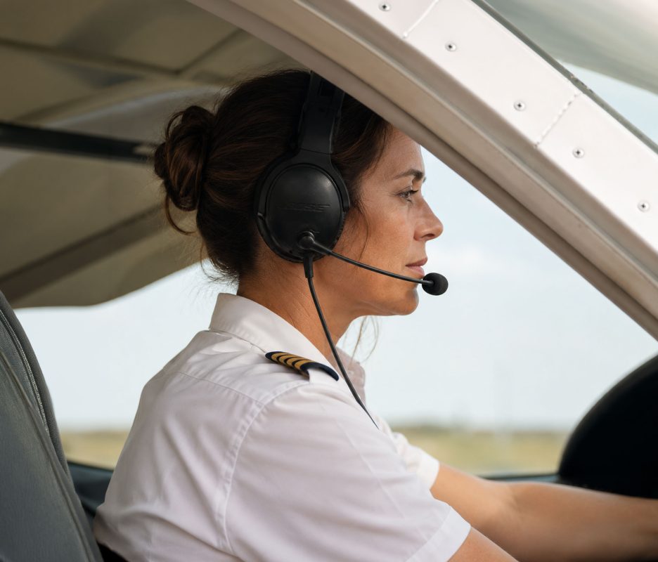 Experienced female pilot seated in the cockpit of a light aircraft, viewed from the side through the open cockpit window. Wearing a white pilot shirt with captain stripes and an aviation headset, she looks ahead with a calm, focused expression while preparing for or conducting a flight. Natural daylight and the aircraft's clean interior emphasize professionalism, confidence, and precision in aviation.