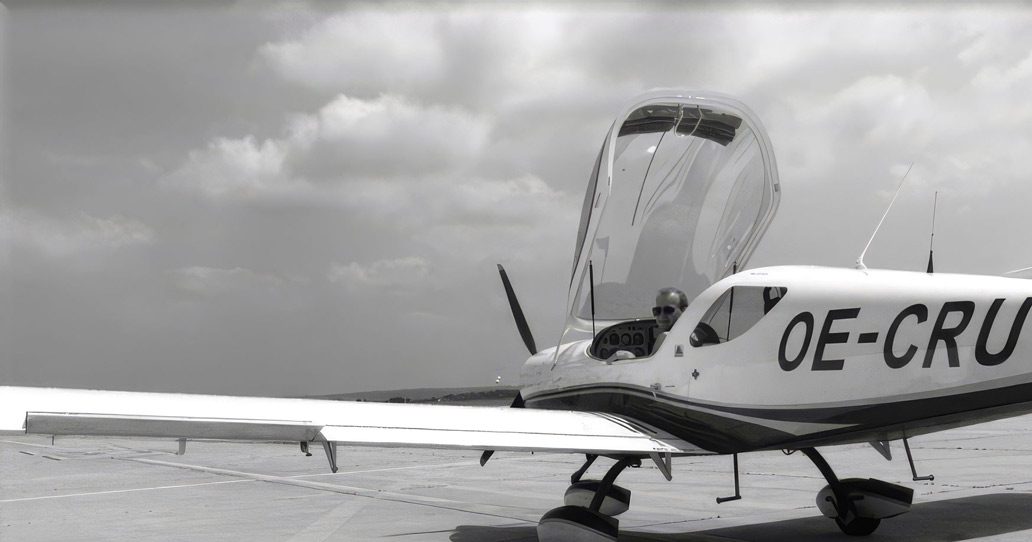 A white single-engine aircraft sits on the tarmac with its cockpit canopy open; a pilot is seated in the cockpit, and clouds are visible in the background.